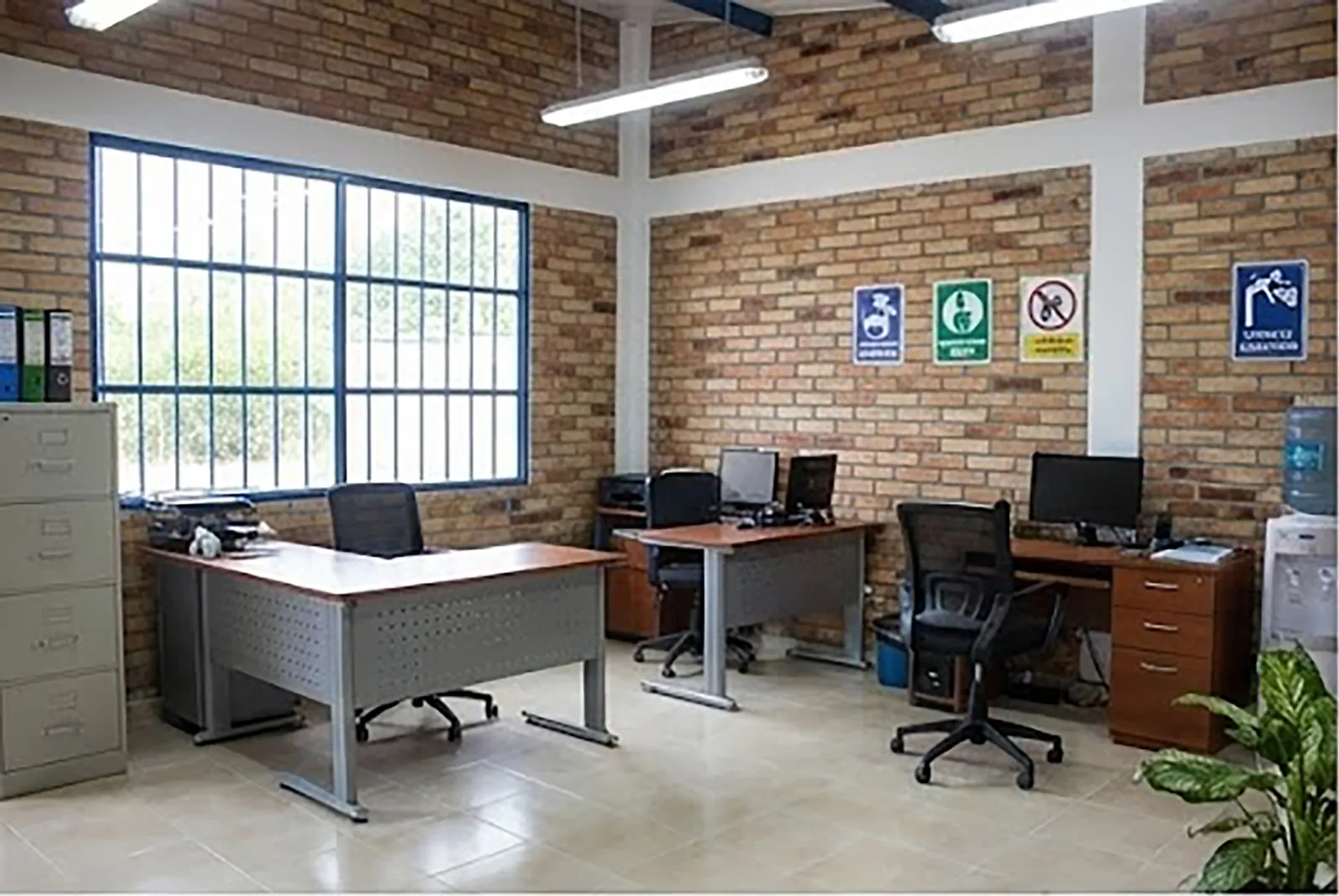 Oficina operativa con puestos de trabajo, computadores, archivador y señalización de seguridad en muros de ladrillo